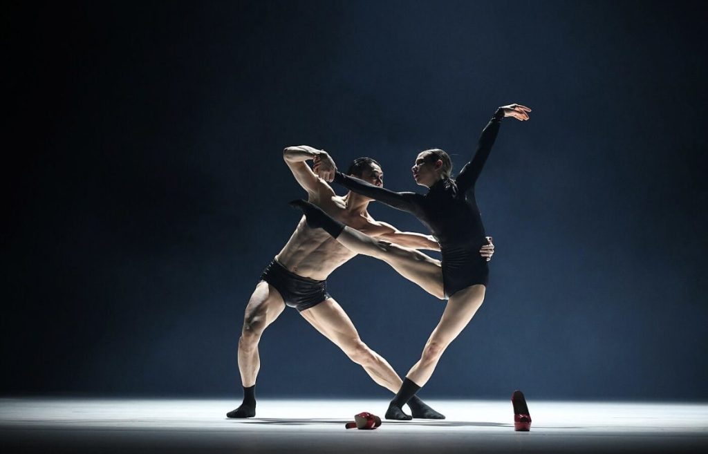 A stage duet in low blue light, a pair of red shoes left on the floor.
