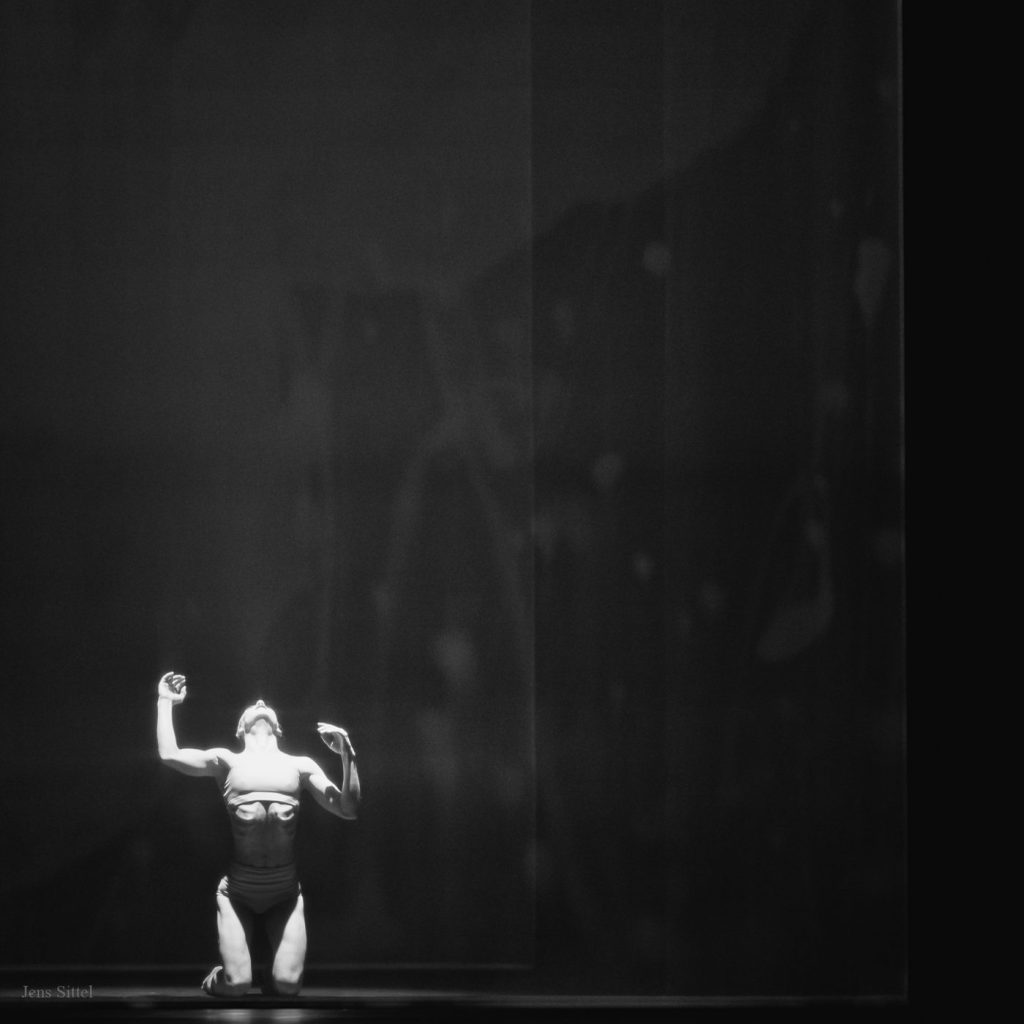 A solo figure kneeling in a narrow shaft of light on a dark stage.